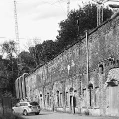 Zwart-witfoto van het fort van Lier vanaf de Fortweg — de lage bakstenen frontgevel met groene begroeiing erbovenop, een wagen op de gezamenlijke parking.