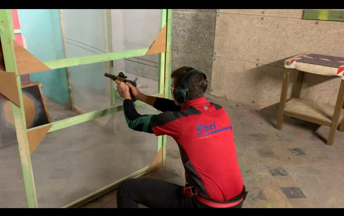 Een lid van Fort Lier in clubpolo schiet geknield een IPSC-parcours, vurend door een schietopening in een barricade.