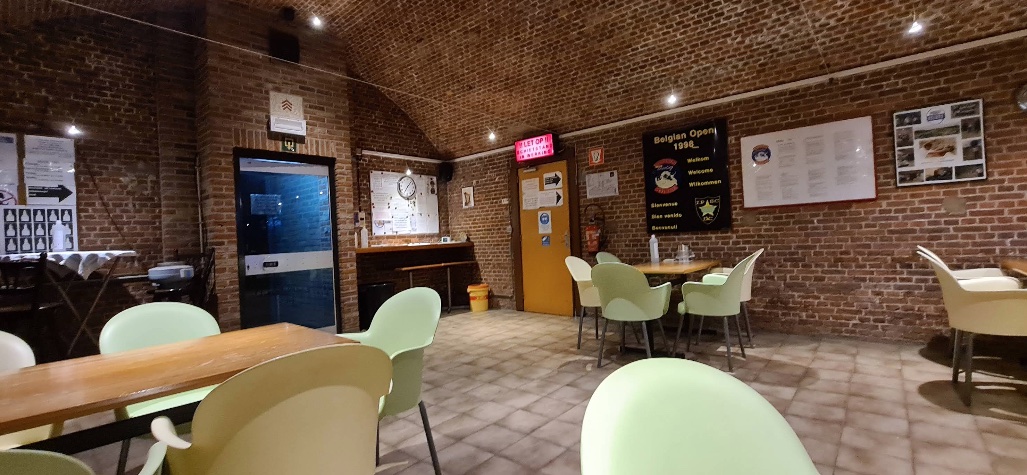 Zithoek van de kantine met tafels en stoelen onder het bakstenen gewelf, met een wedstrijdbanier 'Belgian Open' aan de muur.